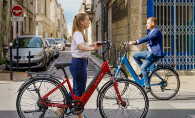 Une femme poussant un vélo MoBY rouge avec, en arrière plan un homme roulant sur un vélo MoBY bleu