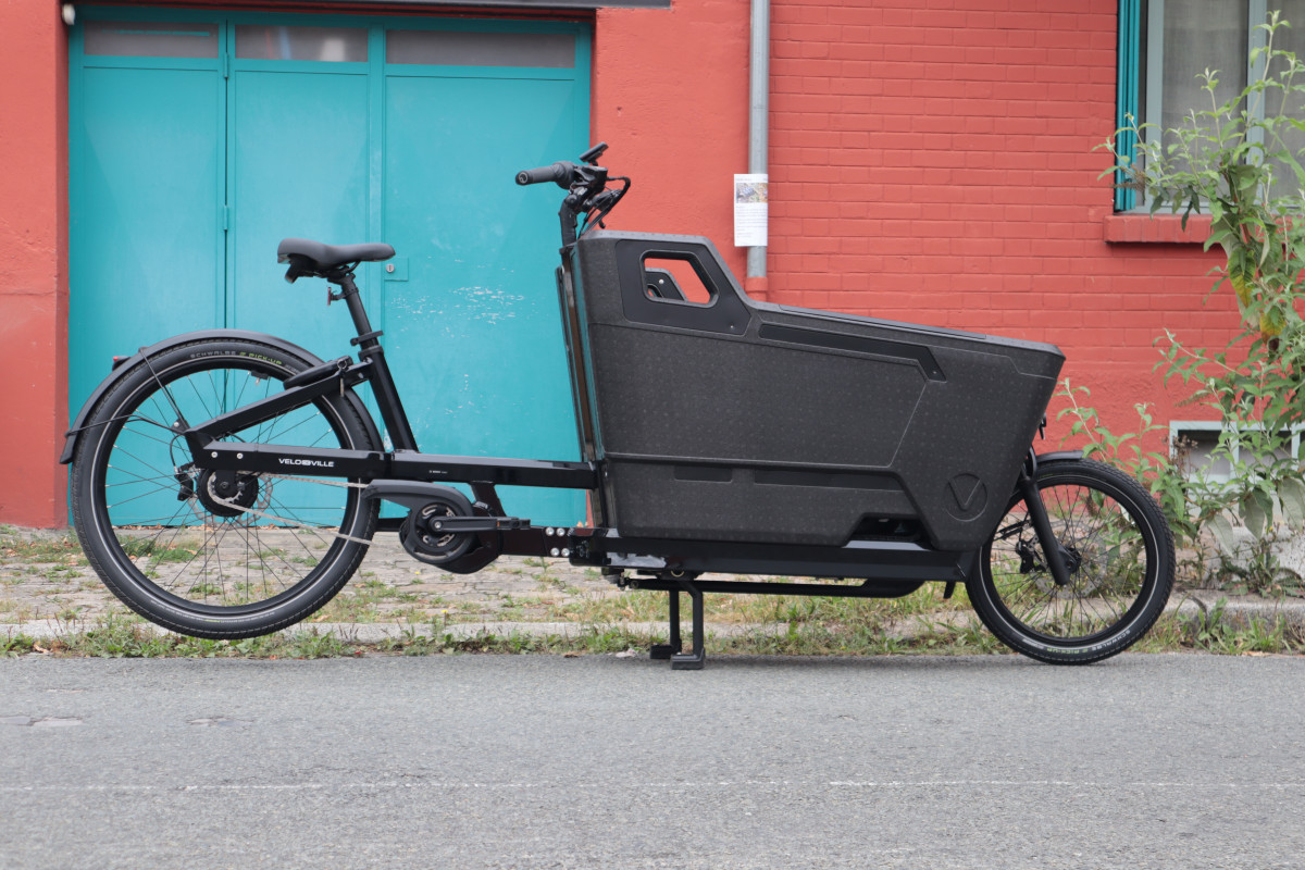 velo cargo FR8 de la marque Velo de Ville. Le vélo tout noir avec sa caisse sur l'avant est posé de profil devant une maison bleu et rouge